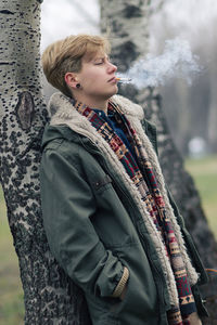 Side view of young woman smoking while standing by tree at park