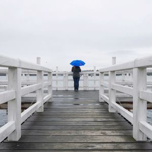 Rear view of woman with blue umbrella standing on pier against sky