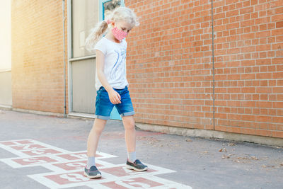 Child girl in face mask playing jumping hopscotch on school yard. new normal.