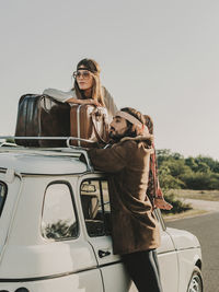 Side view of romantic hippie couple looking away while sitting on old timer automobile with suitcase during trip in nature