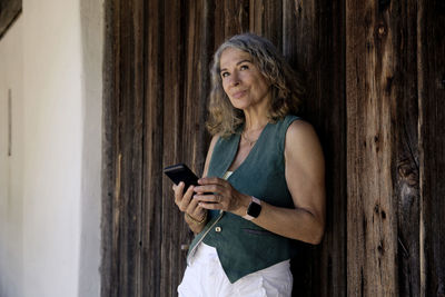 Thoughtful senior woman holding mobile phone leaning on old barn door