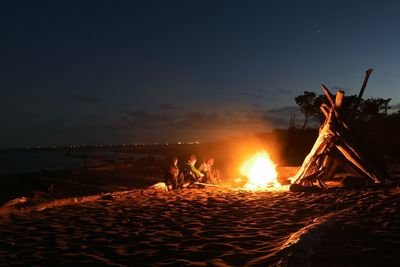Kids sitting by bonfire at beach against sky during night