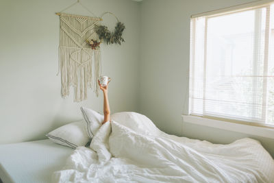 Woman holding coffee mug while wrapped in blanket on bed at home