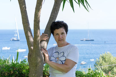 Portrait of smiling woman standing by tree against sea