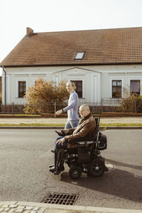 Side view of senior man with disability in motorized wheelchair by young caregiver walking on road during sunny day
