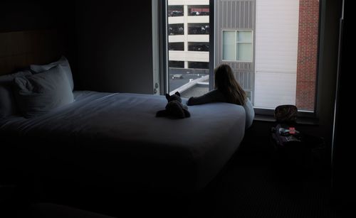 Upset girl sitting by bed and window in hotel room