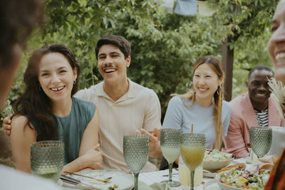 Happy male and female friends talking while sitting together at dining table in back yard