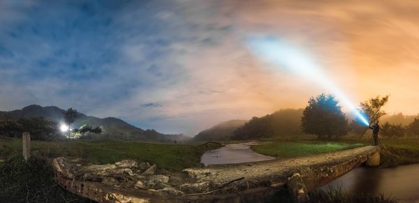 Man with illuminated headlamp standing by river against sky