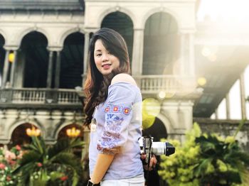 Side view portrait of woman holding camera while standing against historic building