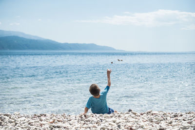 Rear view of boy throwing stones into sea while sitting against sky