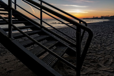 Metallic staircase at beach against sky during sunset