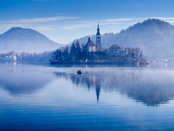 People rowing boat in lake by church and mountains against sky during foggy weather