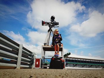 Full length of female operator sitting by camera in stadium against sky