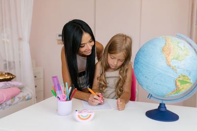 Young woman and her cute little daughter are sitting at a table and having fun doing homework