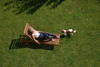 Woman resting on a wooden deckchair on a garden lawn while her dog is playing around