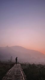Man standing on boardwalk against sky during sunset