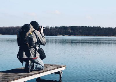 Man photographing while kneeling on pier over lake against sky 