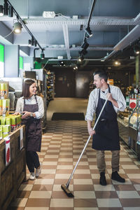 Smiling salesman sweeping floor while looking at colleague in supermarket