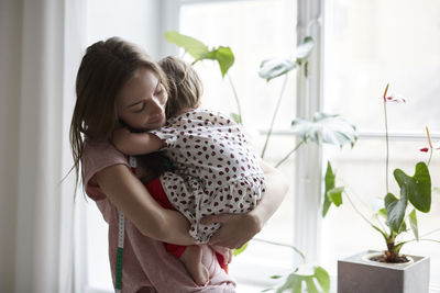 Female fashion designer embracing daughter while standing by window at home