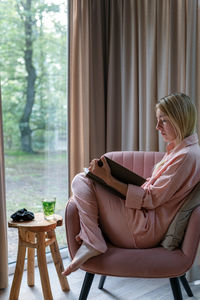A blond woman in pink pajama sits in the pink armchair and reads a book.