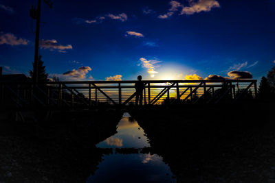 Silhouette boy on bridge over lake against sky during sunset