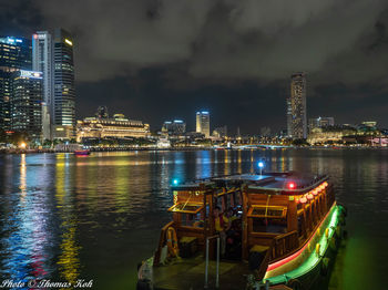 Illuminated modern buildings by river against sky at night