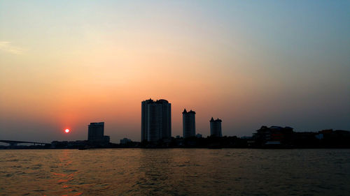 Silhouette buildings by sea against sky during sunset