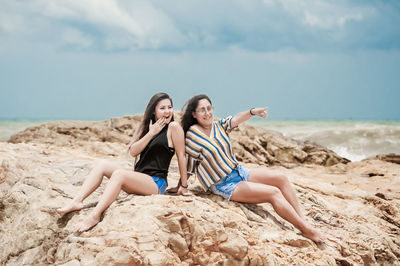 Friends sitting on rock by sea against sky