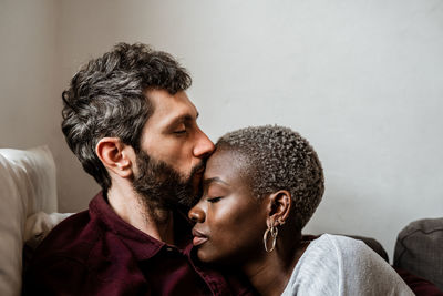 Young bearded man in casual shirt lying on comfortable couch in embrace with african american woman in cozy living room with closed eyes