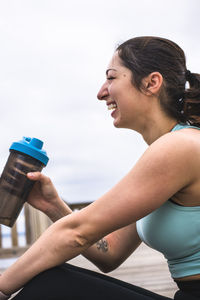 Side view of happy woman holding water bottle while sitting on pier against sky