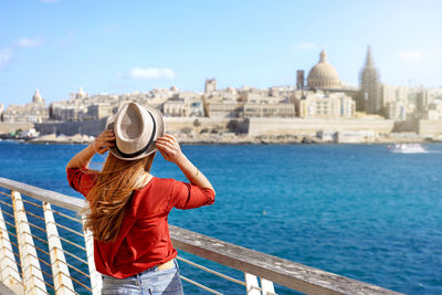 Traveler girl holding hat walking along malta promenade with valletta cityscape on the background.