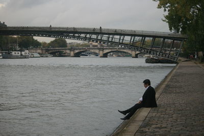 Businessman sitting on footpath by river with arch bridges in background