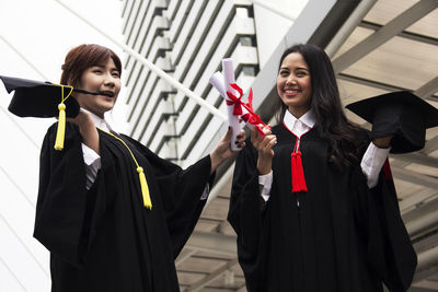 Cheerful young women holding degree certificates standing against sky