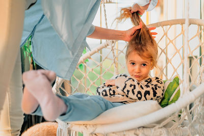 Mother collects hair in a ponytail little daughter in spring at sunset on balcon hammock chair
