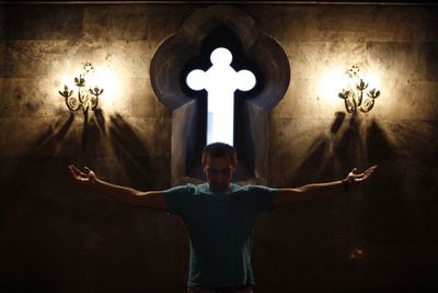 Man with arms outstretched standing against cross in illuminated church