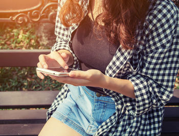 Midsection of woman using smart phone while sitting on bench at park