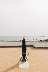 Young woman practicing headstand while doing yoga on rooftop against sky