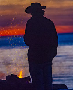 Silhouette man standing by bonfire against sky during sunset