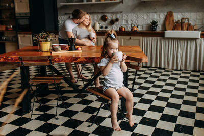 A little girl is holding a mug drinking tea and her parents are kissing from behind in the kitchen