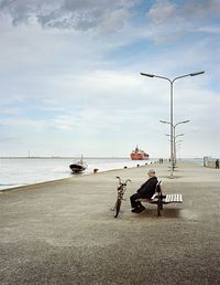 Side view of senior man sitting on bench against sea and cloudy sky