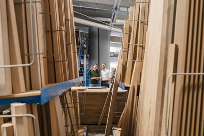 Two carpenters talking in production hall with wooden planks in foreground