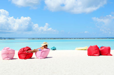 Woman resting on bean bags at beach against sky during sunny day