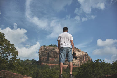 Rear view of man holding camera while standing against sky