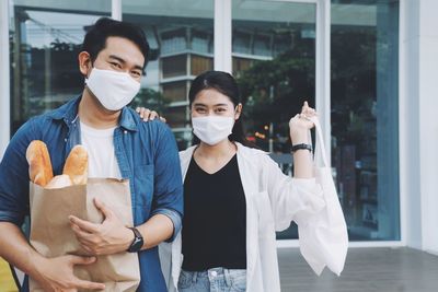 Portrait of smiling couple wearing flu mask holding shopping bag standing outdoors