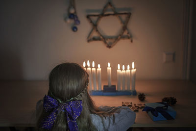 Rear view of girl looking at burning candles against wall at home
