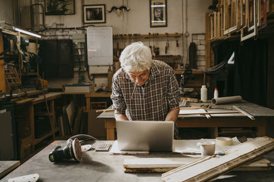 Senior male carpenter holding pencil in mouth while working on laptop at workshop