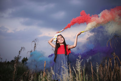 Smiling cute girl holding distress flares while standing against sky at dusk