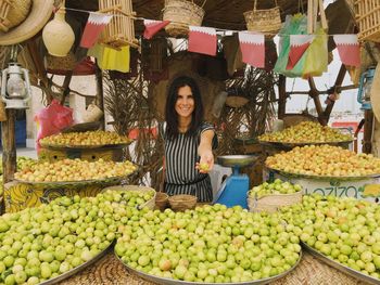 Portrait of smiling woman holding fruits for sale at market stall in doha, qatar.