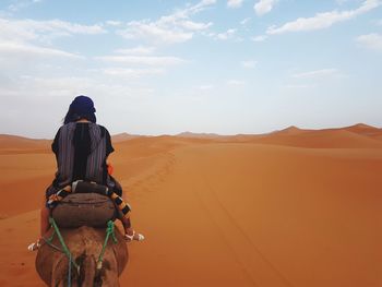 Rear view of woman riding camel at desert against sky