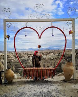 Portrait of woman gesturing while standing by heart shape swing against sky